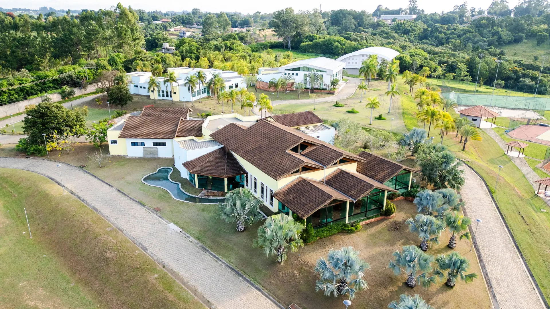 Vista aérea aproximada do Spa Recovery, com o prédio principal de telhado marrom, o ginásio e as palmeiras